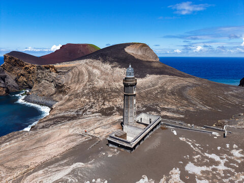 Aerial view of a stark, volcanic landscape dominated by the Capelinhos Lighthouse against the backdrop of a blue sea, Capelo, Acores, Portugal.