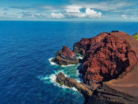Aerial view of the rugged, rust-colored cliffs meeting the deep blue sea, a stark contrast under the vast sky, Capelo, Acores, Portugal.