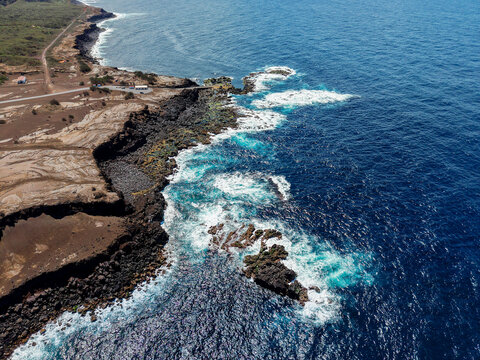 Aerial view of the rugged coastline where the dark volcanic cliffs meet the foamy, turquoise sea, Capelo, Acores, Portugal.