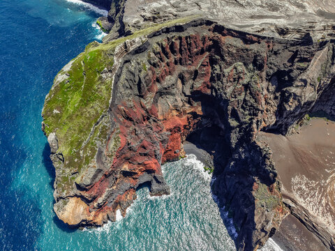 Aerial view of the rugged Capelinhos Volcano coastline, where stark black volcanic rock meets vibrant green vegetation and turquoise waters, Capelo, Acores, Portugal.