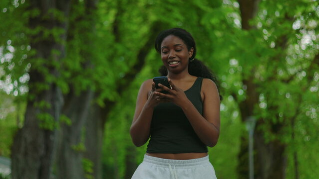 African American woman walking forward while using smartphone and smiling at screen creating positive and outdoor lifestyle moment in green environment with tracking camera movement