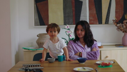 Fototapeta premium Mother watching toddler refuse breakfast while sitting at table with crossed arms, child playing with spoon and food, candid parenting moment showing patience and everyday family dynamic