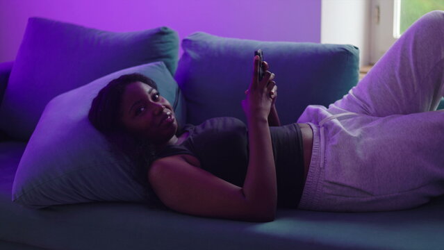African American woman lying on couch using smartphone then looking toward camera briefly before returning to screen, casual interaction and moment of awareness in relaxed home setting