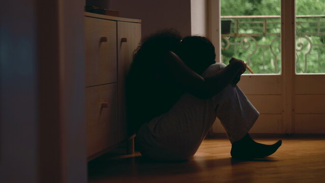 African American woman curled up on floor by window resting head on arms showing emotional collapse exhaustion depression and overwhelming sense of despair in quiet interior