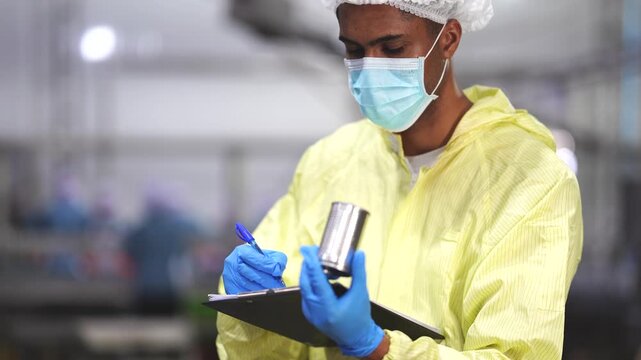 A focused industrial inspector in a yellow protective suit and mask recording quality control data while overseeing an automated production line in a manufacturing plant.