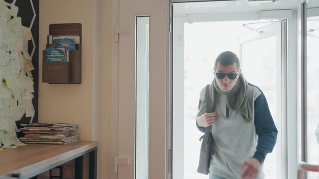 Young man entering cafe through door, wearing sunglasses and scarf, carrying satchel casual customer approaches counter, warm interior with stools and decorative lights, confident relaxed stride
