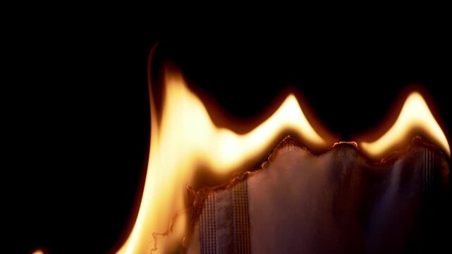 Bright orange flames consume the top edge of a piece of paper against a stark black background. Concept of destruction, intense heat, danger, and the irreversible passage of time. Slow motion.