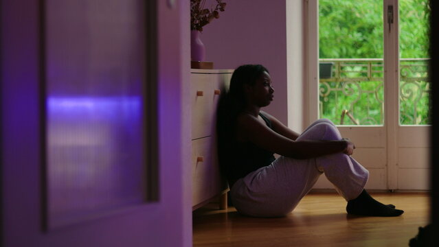 African American woman wide shot sitting on floor by window leaning against cabinet in still posture showing introspection worry and aimless reflection in quiet home space