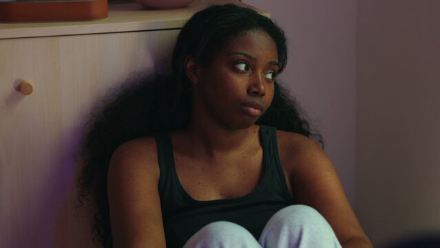 African American woman sitting on floor leaning against cabinet looking aside with distant expression suggesting dissatisfaction contemplation and emotional disengagement in home interior