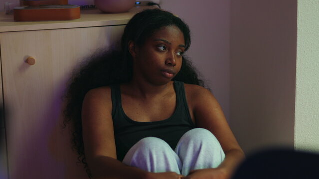 African American woman sitting on floor leaning against cabinet looking aside with distant expression suggesting dissatisfaction contemplation and emotional disengagement in home interior