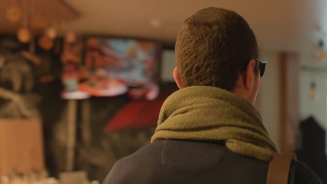Man arriving at cozy cafe entrance, back view wearing scarf, checking mirror reflection, warm lights and soft bokeh create introspective urban mood, subtle film grain, waiting at counter.