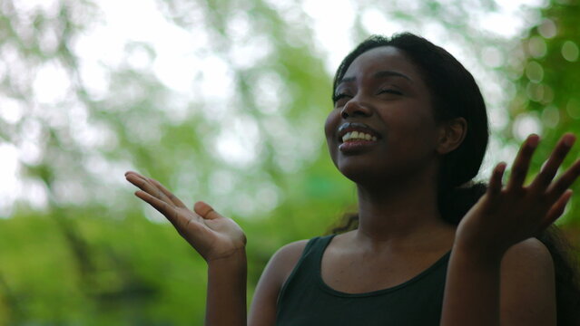African American woman standing outdoors raising hands and looking upward before bringing them together in prayer creating emotional and spiritual moment of hope and faith in natural environment