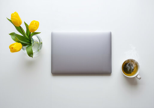 Flat lay of closed laptop with yellow tulips and hot tea on white desk
