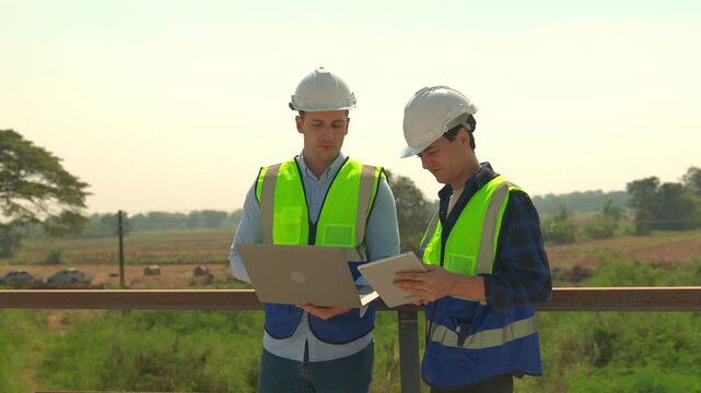 Construction engineer team in hard hat and safety vest using laptop and tablet at outdoor site for field inspection and project coordination in daylight