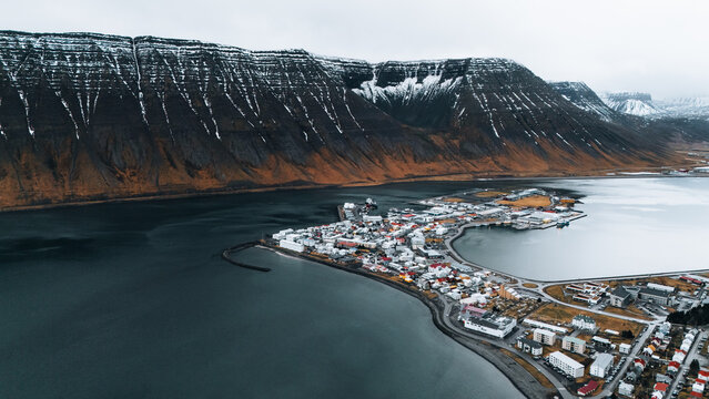 Aerial view of Isafjordur nestled between still waters and rugged mountains, a tapestry of red-roofed buildings contrasting with the snow-dusted peaks, Isafjordur, Iceland.