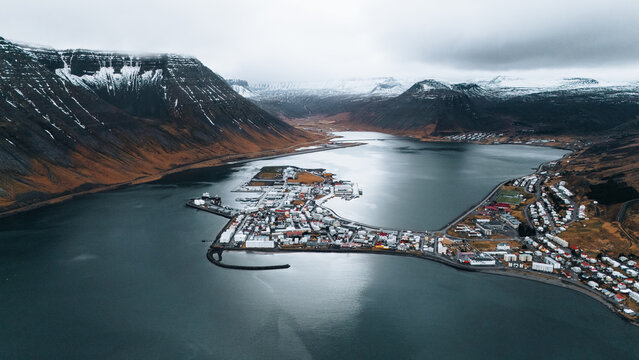 Aerial view of a quaint town nestled between dark mountains capped with snow and the still, reflective waters of the fjord, Isafjordur, Iceland.