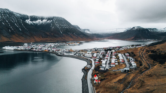 Aerial view of stark contrasts where dark waters meet the rugged coastline, nestled beneath snow-dusted mountains, Isafjordur, Iceland.