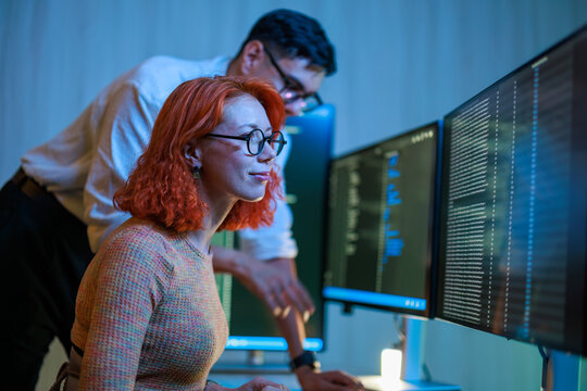 A focused male and female software developer team wearing glasses collaborating late at night, analyzing complex computer code on multiple dual monitors in a dark modern office environment.