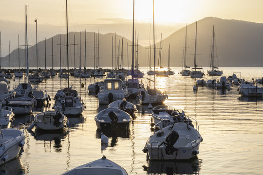 Lerici, Italy - 08 March 2026: View of a harbor filled with boats, their masts reaching towards the sky as the sun sets behind the distant mountains, painting the water in warm, golden hues.