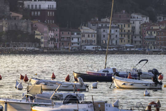 Lerici, Italy - 08 March 2026: View of small boats bobbing gently in the harbor, reflecting the golden light of the setting sun against the backdrop of the pastel-colored buildings..