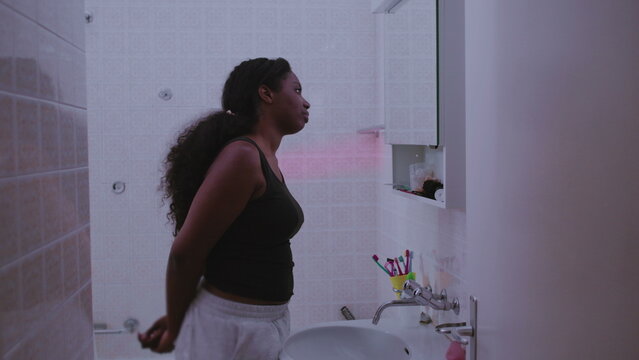 African American woman leaning toward bathroom mirror inspecting skin closely with concerned expression, focusing on facial imperfections, self image, and quiet dissatisfaction with appearance