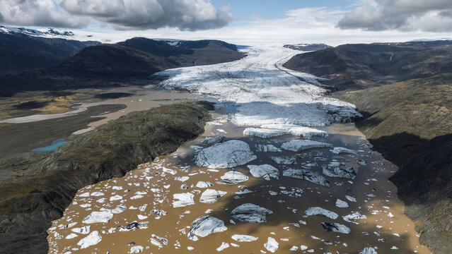 Aerial view of a glacier meeting a silt-laden river, where icebergs drift, contrasting sharply with the rugged, dark terrain beneath a cloudy sky, Hoffellsjokull, Iceland.