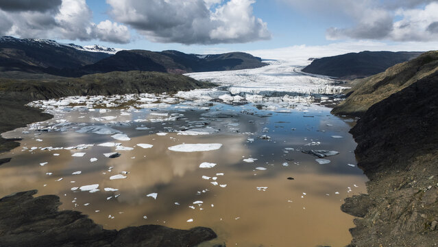 Aerial view of glacial ice drifts serenely on the murky waters, framed by rugged mountains under a textured sky, Hoffellsjokull, Iceland.
