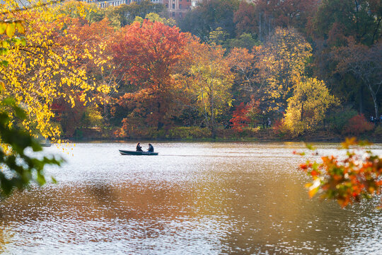 Manhattan, United States - 08 November 2025: View of a tranquil lake reflecting the vibrant autumn foliage, with a small boat gliding across its shimmering surface.