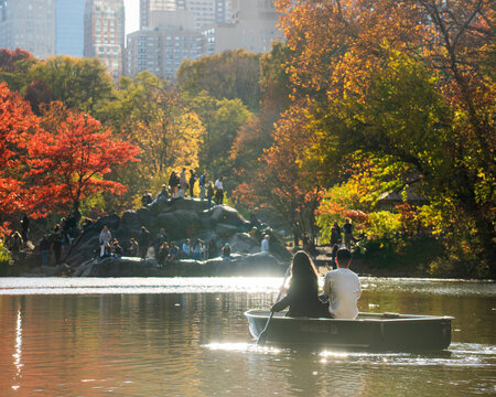 Manhattan, United States - 08 November 2025: View of a serene lake reflecting the vibrant autumn foliage of Central Park, with people gathered on rocks and a couple gliding by in a rowboat.