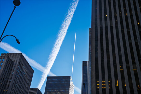 Manhattan, United States - 03 December 2025: View of towering skyscrapers piercing a bright blue sky, crisscrossed by vapor trails, creating an urban tapestry of steel and sky.