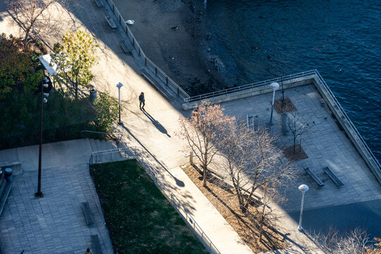 Manhattan, United States - 01 December 2025: View of a lone figure strolling along the East River waterfront, bisected by sharp sunlight and shadows, where the city meets the tranquil water's edge.