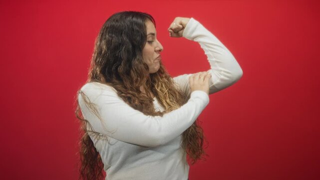 Woman flexes right bicep and presses hand to upper arm wearing white long sleeve top against red studio backdrop while looking at muscle with a smirk; confidence.