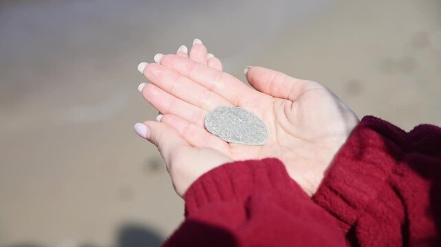 Hands holding small smooth grey stone outdoors with soft natural light on blurred sandy background