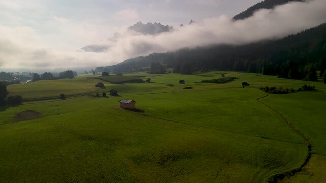 A typical wooden cabin stands in a meadow in the Bolzano region of South Tyrol. In the background rise the majestic mountains of the Sexten Dolomites, shrouded in clouds. drone perspective.