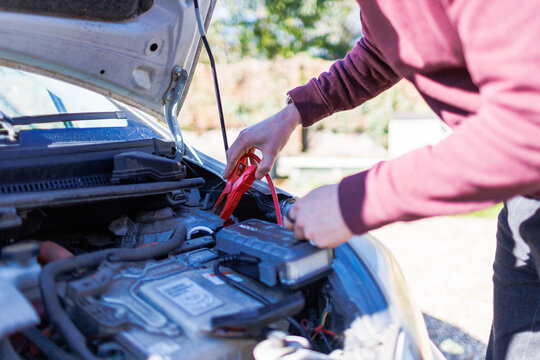 Young man using jumper cables to boost a car battery under the hood on a sunny day, roadside emergency assistance concept
