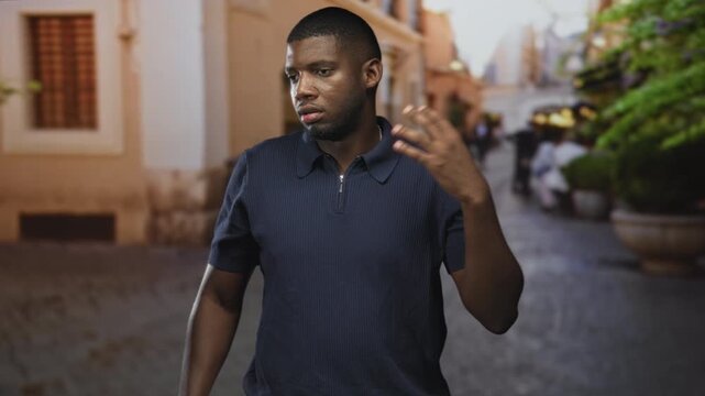 Young man adjusting polo collar with raised hand while glancing aside on a sunlit cobbled street with potted planters; thoughtful introspection.