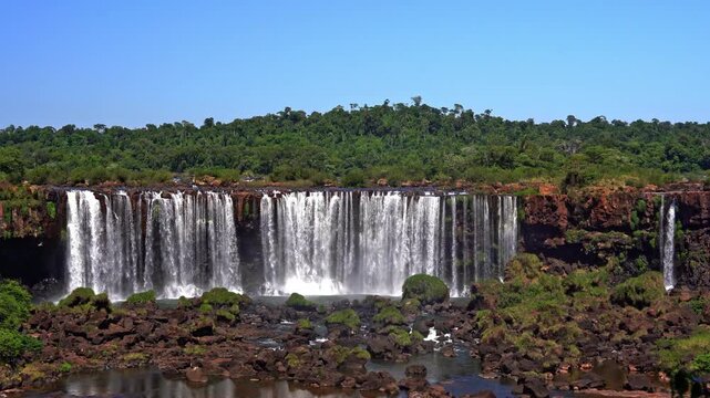 Panoramic view of the majestic Iguazu Falls in Argentina, powerful waterfalls cascading over rocky cliffs surrounded by lush tropical jungle under a clear blue sky.