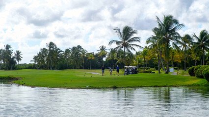 Obraz premium Group of golfers preparing to tee off on a lush green golf course surrounded by palm trees and a calm water body under a partly cloudy sky