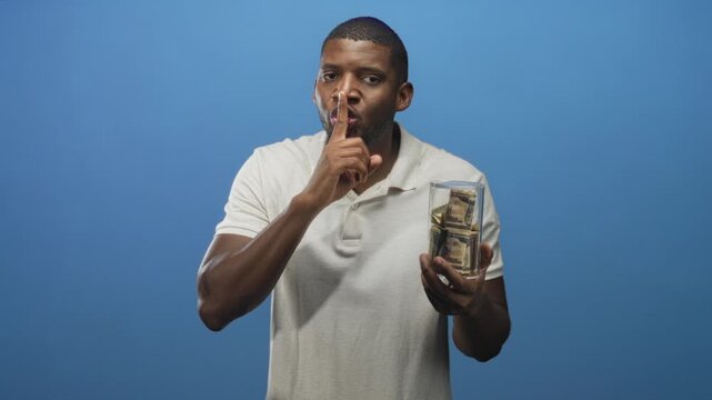 Young man holds glass jar of bundled dollar bills and presses finger to lips for silence in studio; secrecy.