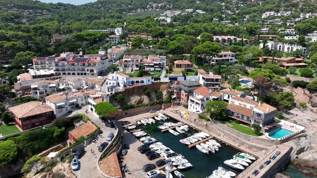 Aerial view of Costa Brava Spain with luxury white and terracotta houses perched on steep cliff edge overlooking green forested hills under bright blue sky