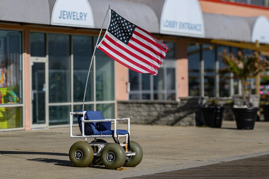 A beach wheelchair, a specialized mobility device designed for traversing sand and uneven terrain.  The American Flag waves in the wind on the Boardwalk at Ocean City MD.