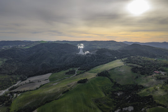 Aerial view of a geothermal power plant cooling tower emitting steam amidst the rolling green hills and forested landscape under a hazy sunset sky in Pomarance, Tuscany, Italy.