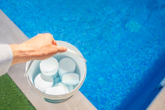 Top view of a person holding a bucket filled with chlorine tablets near a swimming pool, used for water sanitation and maintenance - pool maintenance concept