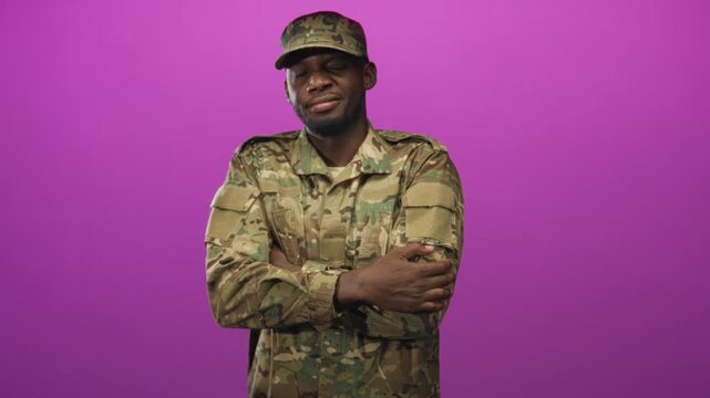 Man in camouflage uniform and cap standing with arms crossed and a slight wink, wearing military jacket in studio; pride duty.