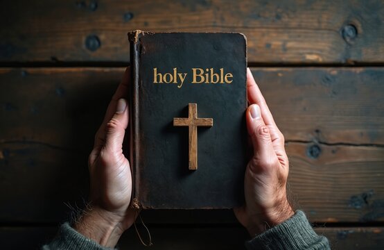 Hands hold old holy Bible with wooden cross on dark rustic table. Close-up view shows texture and faith symbol. Religious book reading and prayer time. Spiritual connection and belief.