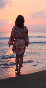 Happy woman is walking on the sandy beach in a red dress against sunrise. Slow motion