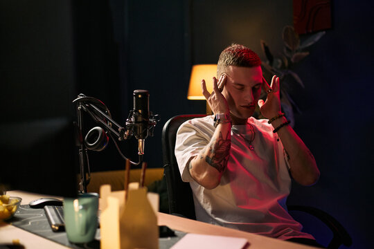 Young adult Caucasian man streaming podcast, touching temples while focusing beside microphone and headphones. Ideal for creator economy, live content, burnout, digital work themes