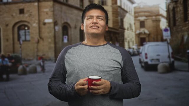 Man holding red mug of coffee with both hands, smiling while looking at camera on a cobblestone street in an old town square; contentment travel pause.