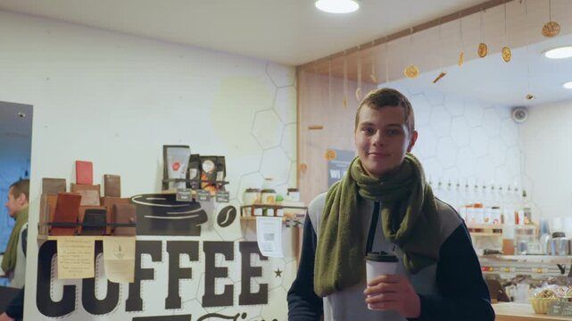 Bustling coffee shop counter with takeaway cup and neon sign showing young man ordering then settling to type, scarf and togo cup in foreground, pastries on display, barista serving, freelancer