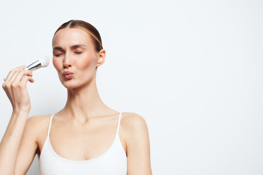 Young woman with clean skin applies natural deodorant stick on underarm wearing white tank top isolated on white background with closed eyes and relaxed facial expression.